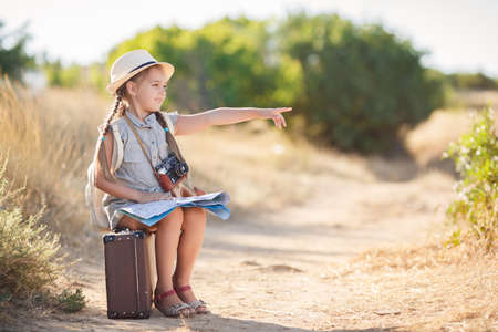 Cute little girl 6 years old, long hair braided into two pigtails, dressed in a summer Onesie blue color, on his head wearing a sun hat, behind wears light leather backpack, sitting on the old suitcase among the back roads, exploring the map of the areaの写真素材