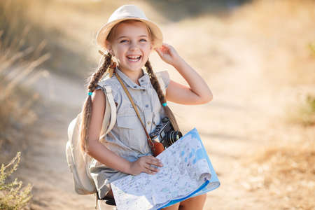 Cute little girl 6 years old, long hair braided into two pigtails, dressed in a summer Onesie blue color, on his head wearing a sun hat, behind wears light leather backpack, sitting on the old suitcase among the back roads, exploring the map of the areaの写真素材