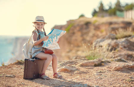 Cute little girl 6 years old, long hair braided into two pigtails, dressed in a summer Onesie blue color, on his head wearing a sun hat, behind wears light leather backpack, sitting on the old suitcase among the back roads, exploring the map of the areaの写真素材