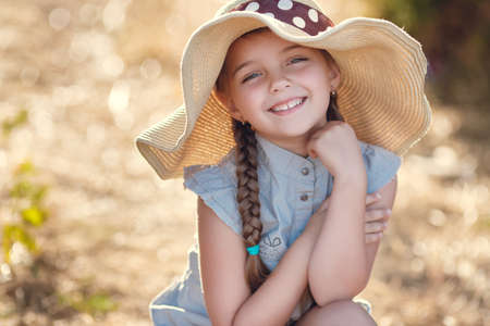 Cute little girl 6 years old, hair braided in two braids, on his head a large straw hat, wearing overalls in the summer gray, in the ears of small earrings, grey eyes and a cute smile posing outdoors in the summerの写真素材