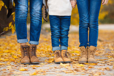 Happy young family, father, mother and little daughter in the autumn Park, all three dressed in blue jeans and brown shoes, standing next to each other on the yellow of autumn leaves the alley of the Park, hold each other's hands, pay attention to the feeの写真素材