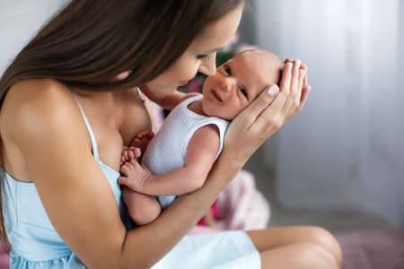 Happy young brunette woman with long straight hair, dressed in a white sundress, spends time with her newborn son in the bedroom, sitting on the floor on a pink fluffy rug, the mother loves the child lying on her lap, dressed in white coverallの写真素材