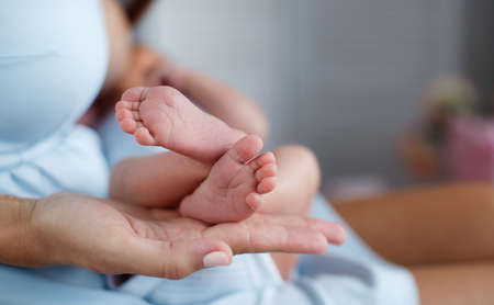 Happy woman, brunette, white mother of pearl nail Polish, gently hold in their palms a little pink feet newborn baby photo on a pink background in the bedroomの写真素材