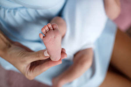 Happy woman, brunette, white mother of pearl nail Polish, gently hold in their palms a little pink feet newborn baby photo on a pink background in the bedroomの写真素材
