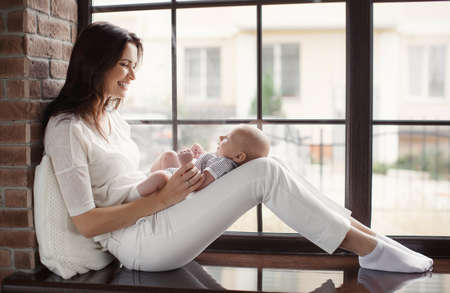 A beautiful woman, a young mother, brunette with long hair, dressed in a white T-shirt and white pants on her lap her newborn baby, dressed in a striped t-shirt sitting on a brown varnished window sill near the window, wearing a ring on his right handの写真素材