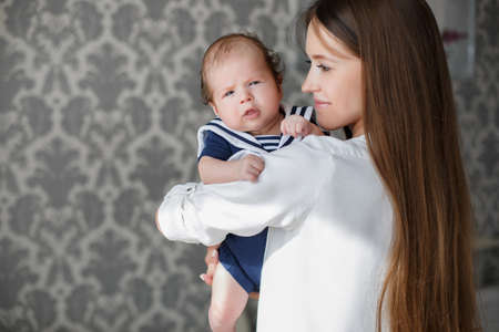 Portrait of mother and child, beautiful mother, brunette with long straight hair, dressed in a white shirt, is holding his newborn baby boy with curly hair, dressed in a dark blue jumpsuit sailor, standing together on a gray background Wallpaperの写真素材