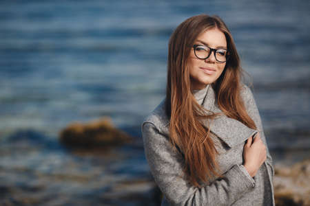 Stylish young woman model looks with long blond hair, wears black-rimmed glasses, slim figure, light makeup, dressed in a grey turtleneck and a grey coat posing on the street alone on a deserted beach on a blue sea backgroundの写真素材