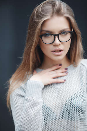 Young beautiful woman with model looks, wears black-rimmed glasses, beautiful makeup, pale pink lipstick, long red hair, gray eyes, wearing a light knitted sweater posing in Studio on dark gray backgroundの写真素材