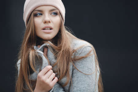 Young beautiful woman with model looks, beautiful makeup, pale pink lipstick, long red hair, big black eye lashes, blue eyes, wearing a light gray coat and a pink sports hat posing on grey background in the city in the fallの写真素材