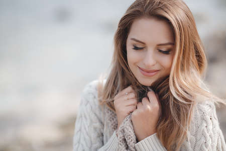 Spring portrait beautiful woman with blond, long, straight hair, big black eyelashes, drooping eyelids, light makeup, pink lipstick, wearing jewelry, wearing a knitted sweater is white with a large beige collar, posing outdoors in early spring.の写真素材