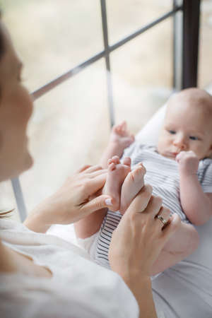 A beautiful woman, a young mother, brunette with long hair, dressed in a white T-shirt and white pants on her lap her newborn baby, dressed in a striped t-shirt sitting on a brown varnished window sill near the windowの写真素材