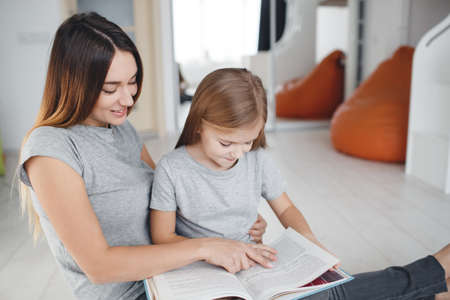 Young woman brunette with long straight hair, wearing a light gray t-shirt and grey jeans sitting on white floor with his daughter, a girl with blond long hair, wearing a light gray t-shirt, mother and daughter reading a bookの写真素材
