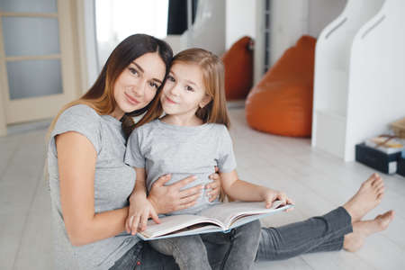 Young woman brunette with long straight hair, wearing a light gray t-shirt and grey jeans sitting on white floor with his daughter, a girl with blond long hair, wearing a light gray t-shirt, mother and daughter reading a bookの写真素材