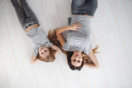 Young beautiful mother and her little daughter lie on your back on a white wooden floor, both dressed in light gray T-shirt and gray jeans, both beautiful brown eyes and long straight hair chestnut color, pleasant smileの写真素材