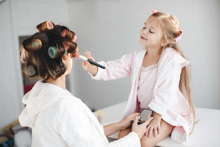 A little girl with long blond hair and her young mother, brunette, dressed in dressing gowns, are in a bright room, the girl sitting on a white table, using cosmetic brushes mother and daughter are doing makeup together, hair wound on large curlers pinkの写真素材