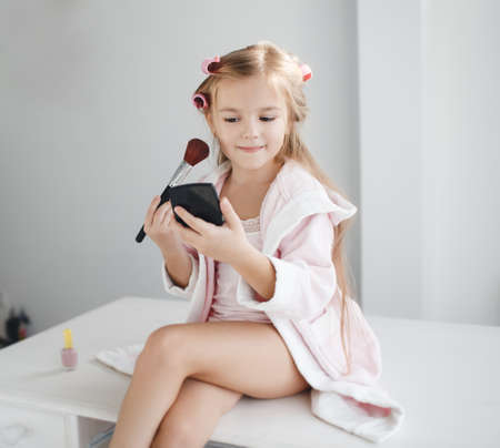 Little girl with long blond hair and brown eyes, wind the hair on the big pink curlers, holding in his hand a black powder compact and a large brush for makeup, sitting on a white table in front of the window, wearing a pink T- shirt and dressing gownの写真素材