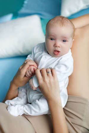 Funny baby with gray eyes and small, bright, fluffy hair, open mouth, dressed in white overalls, playing with his mother, sit on her stomach, the mother lies on the blue bed, bending the knees, wearing a dress of beige, wearing a ring on handの写真素材