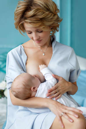 Young happy mother in white house dress with short sleeves, sitting on the bed, tenderly holding his newborn son, the baby sucks mother's breast and falls asleep, the child is dressed in white overalls, a small stick is lying on the mother's breastの写真素材