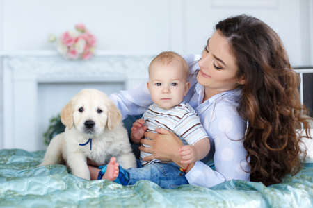 Little boy with short blond hair, barefoot, sitting on the bed on a turquoise blanket with her mother, a beautiful woman with long curly hair, black hair, wearing a white shirt and blue jeans, along with them - a pet, puppy golden retriever , happy familyの写真素材