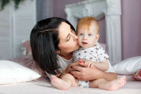 Funny kid with red hair and blue eyes, dressed in a white t-shirt with grey stars and plays on the bed in the bedroom with his mother, a woman with black hair, dressed in a white t-shirt and blue jeans, the woman's nails painted red nail Polishの写真素材