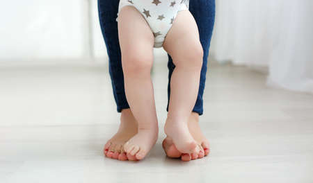 Bare legs of the child, dressed in a white diaper, standing on the bare soles of the feet of mother dressed in blue jeans, baby feet are between mothers legs, portrait on the background of the home environment.の写真素材