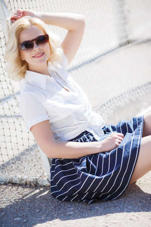 A beautiful young woman, blonde, wearing sunglasses, dressed in a dark blue sundress with white stripes off the shoulder white shirt posing outdoors in the summer, standing on the Playground near the football gateの写真素材