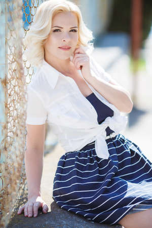 A beautiful young woman, blonde with brown eyes, dressed in a dark blue sundress with white stripes off the shoulder white shirt posing outdoors in the summer, standing on the Playground near the football gateの写真素材
