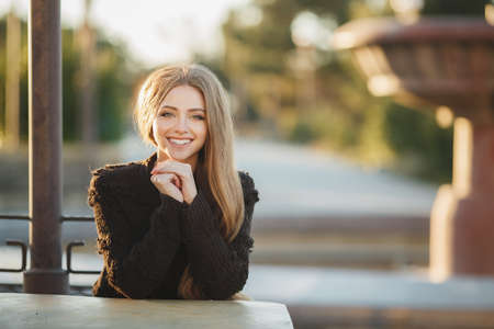 Young, beautiful, attractive woman, brunette with long straight hair and gray eyes, a beautiful smile and straight white teeth, dressed in a black knitted pullover, sitting at a table in the street cafe, posing against a background of an urban landscape.の写真素材
