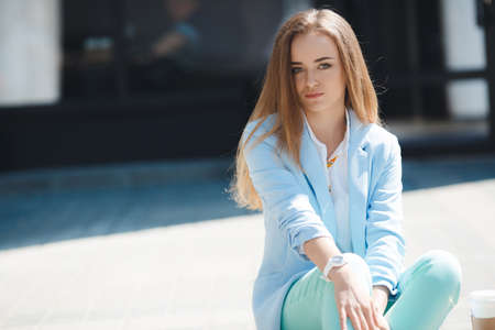 Beautiful young, stylish woman with gray-green eyes, long straight blond hair, sitting on the sidewalk near his office in the open air in summer, dressed in a white shirt and pants suit blue colorの写真素材