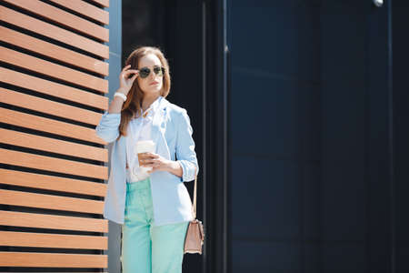 A stylish, young, beautiful woman in a blue trouser suit, white shirt, with a beige handbag, light long straight hair, light make-up, sunglasses, posing alone near her office outdoors in the summer with a paper cup of cappuccinoの写真素材