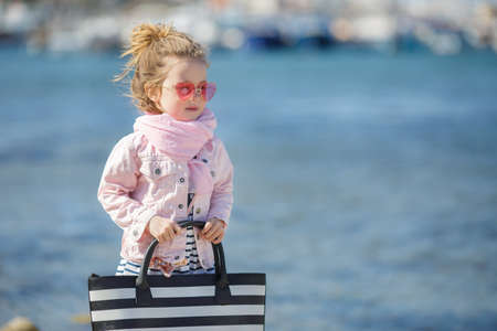 Beautiful little girl with blond hair, wearing pink glasses in the shape of hearts, big beach black and white striped bag, striped skirt, pink jacket, pink scarf, spending time alone outdoors on the beach in the afternoon, posing on blue ocean backgroundの写真素材