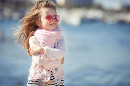Beautiful little girl with blond hair, wearing pink glasses in the shape of hearts, striped skirt, pink jacket, pink scarf, spending time alone outdoors on the beach in the afternoon, posing on blue ocean backgroundの写真素材