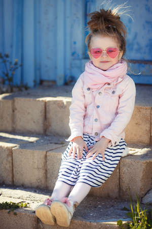 Beautiful little girl with blond hair, wearing pink glasses in the shape of hearts, striped skirt, pink jacket, pink scarf, spending time alone outdoors in the village, sitting on the steps on a blue backgroundの写真素材