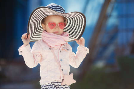Beautiful little girl with blond hair, wearing glasses with pink glasses in the shape of hearts, striped hat with large fields, striped skirt, pink jacket, pink scarf, spending time alone outdoors in the afternoon, posing on blue backgroundの写真素材