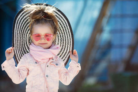 Beautiful little girl with blond hair, wearing glasses with pink glasses in the shape of hearts, striped hat with large fields, striped skirt, pink jacket, pink scarf, spending time alone outdoors in the afternoon, posing on blue backgroundの写真素材