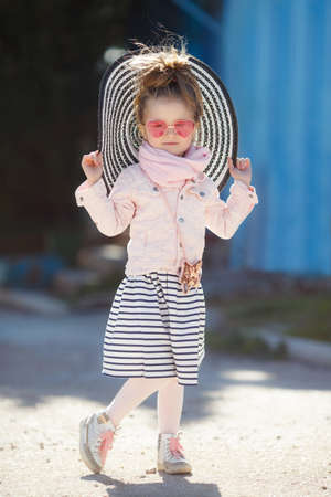 Beautiful little girl with blond hair, wearing glasses with pink glasses in the shape of hearts, striped hat with large fields, striped skirt, pink jacket, pink scarf, spending time alone outdoors in the afternoon, posing on blue backgroundの写真素材