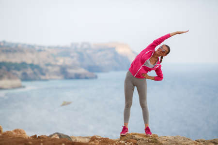 Athletic woman with a sweet smile, brunette, hair in a braid, full lips, wearing a gray t-shirt and gray sweat pants, pink sneakers and a pink sports jacket, wearing dark sunglasses, performing warm-up exercises on the rocky shore The blue oceanの写真素材