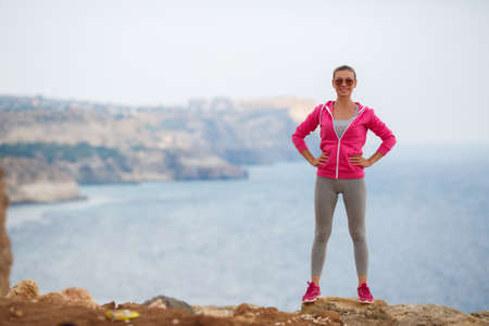Athletic woman with a sweet smile, brunette, hair in a braid, full lips, wearing a gray t-shirt and gray sweat pants, pink sneakers and a pink sports jacket, wearing dark sunglasses, performing warm-up exercises on the rocky shore The blue oceanの写真素材