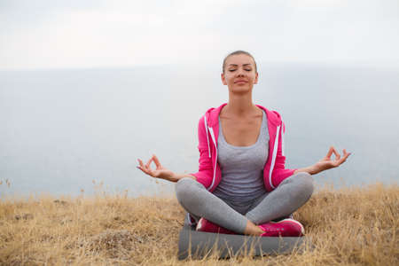 Athletic woman with a sweet smile, brunette, hair in a braid, full lips, wearing a gray t-shirt and gray sweat pants, pink sneakers and a pink sports jacket, performing relaxation exercises, sitting on a rocky beach Lotus positionの写真素材