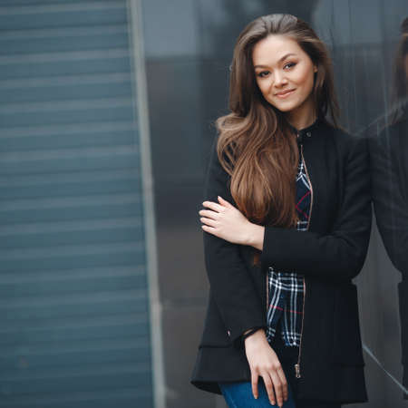 A young leader, a beautiful modern woman, a brunette with long straight hair and brown eyes, long black eyelashes and a sweet smile, is dressed in a black stylish jacket and plaid shirt, posing standing outdoors near the mirror wall of the officeの写真素材