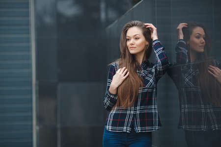 A young leader, a beautiful modern woman, a brunette with long straight hair and brown eyes, light make-up, long black eyelashes and a sweet smile, is dressed in a plaid shirt, posing standing outdoors near the mirror wall of the officeの写真素材