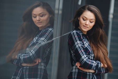 A young leader, a beautiful modern woman, a brunette with long straight hair and brown eyes, light make-up, long black eyelashes and a sweet smile, is dressed in a plaid shirt, posing standing outdoors near the mirror wall of the officeの写真素材