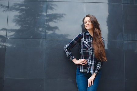 A young leader, a beautiful modern woman, a brunette with long straight hair and brown eyes, light make-up, long black eyelashes and a sweet smile, is dressed in a plaid shirt, posing standing outdoors near the mirror wall of the officeの写真素材