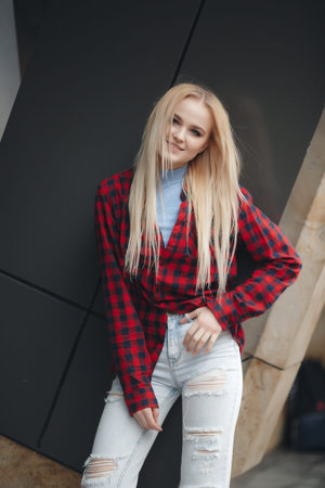 Young attractive woman, blonde with long straight hair and gray eyes, light make-up and cute smile, is dressed in a red checkered shirt with light jeans, posing alone, standing next to a beautiful gray office buildingの写真素材