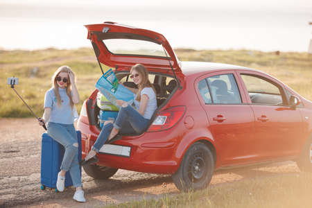 Young, slim, beautiful women, a blonde in a blue dress and a red-haired girl in a red dress, gray eyes, lovely smiles, long black eyelashes, red lipstick and makeup, two best friends are preparing to travel on a red car With suitcases and thingsの写真素材
