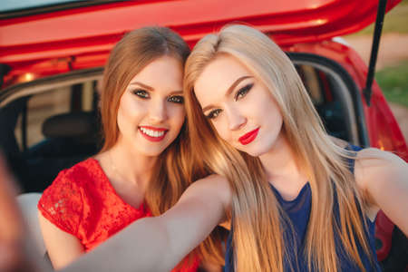 Young, slim, beautiful women, a blonde in a blue dress and a red-haired girl in a red dress, gray eyes, lovely smiles, long black eyelashes, red lipstick and makeup, two best friends are preparing to travel on a red car With suitcases and thingsの写真素材