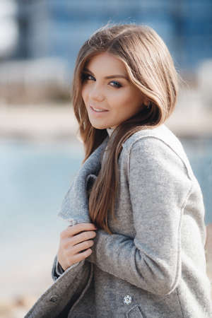 Portrait of a beautiful young woman on an autumn deserted beach, light straight long hair, blue eyes, light make-up, sweet smile, pink lipstick, spends her time alone in the fresh air, walking near the sea, wearing a gray autumn coatの写真素材