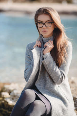 Portrait of a beautiful young woman on an autumn deserted beach, light straight long hair, blue eyes, light make-up, sweet smile, pink lipstick, spends her time alone in the fresh air, walking near the sea, wearing a gray autumn coat , wearing glassesの写真素材