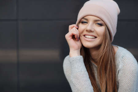 Cute girl with blue eyes, long, straight, light brown hair, beautiful smile, dressed in a knit pullover, gray-blue color, wearing a sports cap pink, light makeup, spends time alone in the city, posing against the backdrop of the gray wall, spring portraitの写真素材