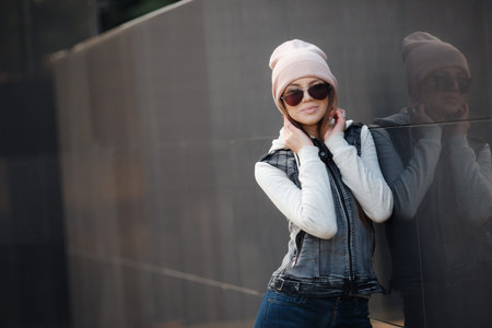 Cute girl in dark glasses, long, straight, light brown hair, beautiful smile, dressed in a knit pullover, gray-blue color, wearing a sports cap pink, light makeup, spends time alone in the city, posing against the backdrop of the gray wall, spring portraiの写真素材
